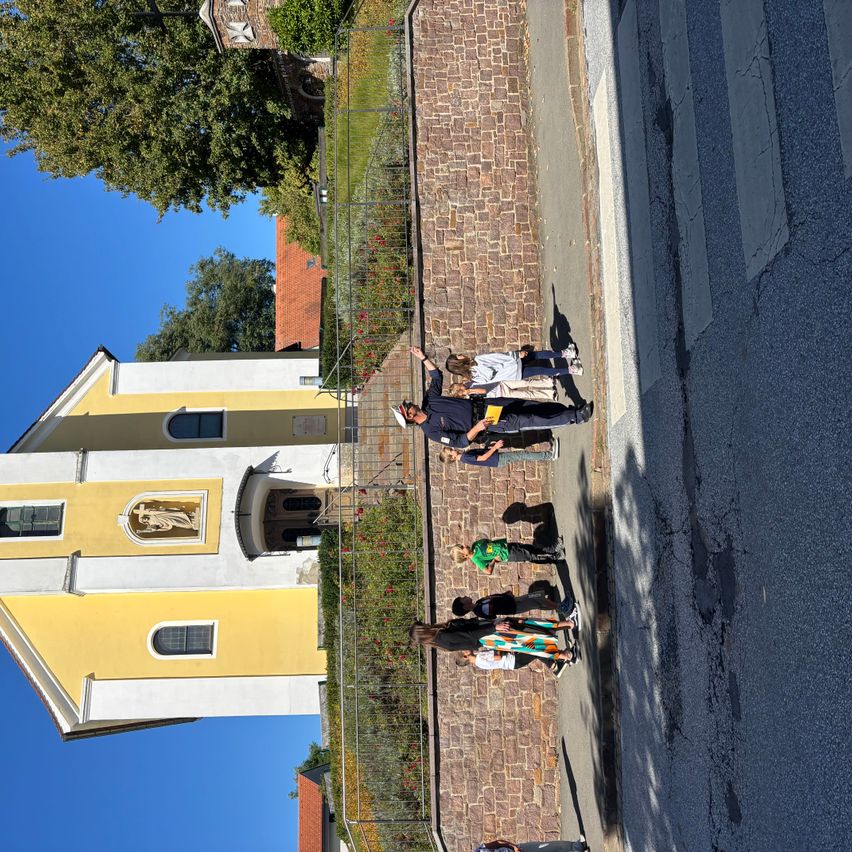 Eine Gruppe von Menschen steht vor einer Kirche mit gelber und weißer Außenfassade. Sie alle blicken auf das Gebäude und scheinen Touristen zu sein.