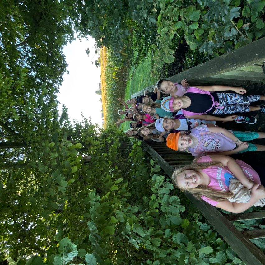 Eine Gruppe von Kindern ist auf einer Holzterrasse, umgeben von Grün, und lächelt in die Kamera. Einige tragen Mützen, und eine hält ein Baby. Bäume und ein Feld sind im Hintergrund.