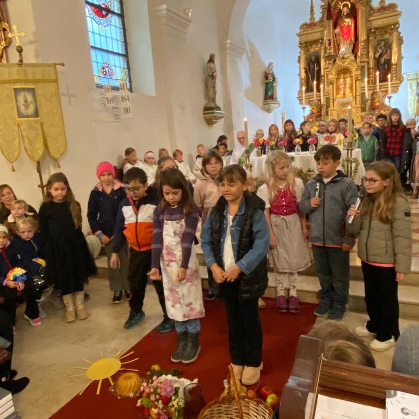 In einer Kirche versammeln sich Kinder für eine Zeremonie, die auf einem roten Teppich stehen. Sie tragen Winterkleidung, und Blumen und eine Sonne sind vor ihnen platziert. Kerzen sind auf dem Altar angezündet.