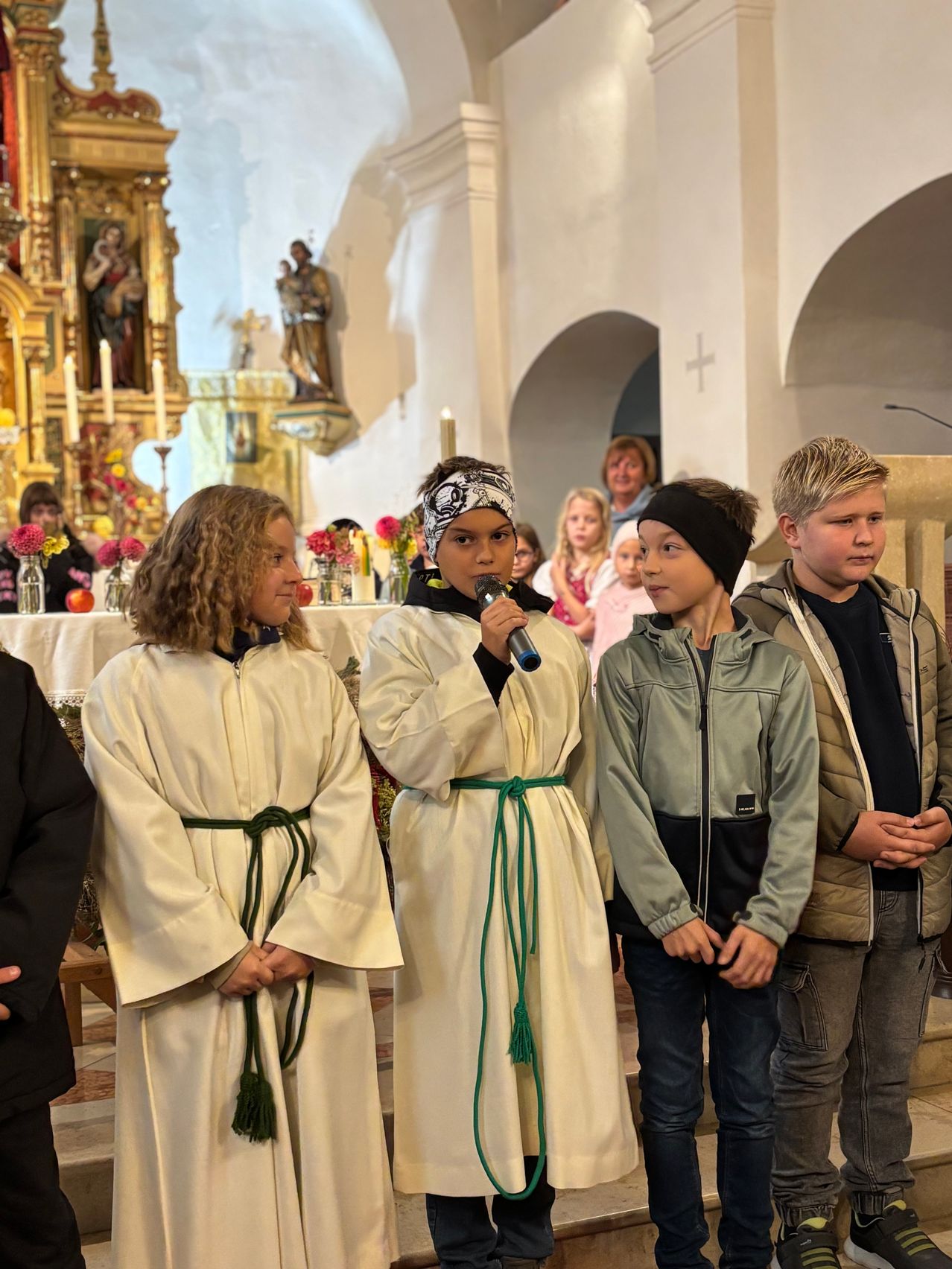 In einer Kirche stehen drei Kinder in weißen Gewändern und eines in Freizeitkleidung vor einem Altar mit Blumen und Kerzen.