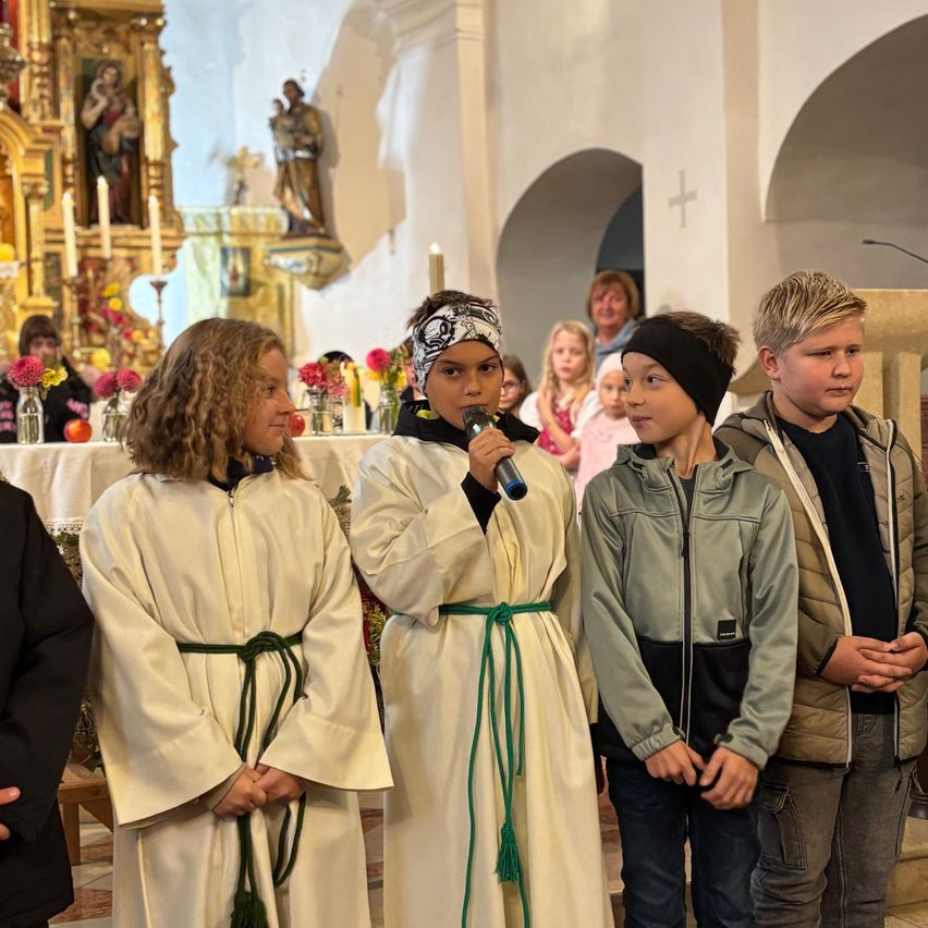 In einer Kirche stehen drei Kinder in weißen Gewändern und eines in Freizeitkleidung vor einem Altar mit Blumen und Kerzen.