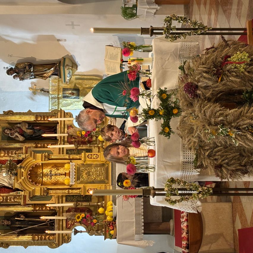 Ein Priester ordnet Blumen vor einem Altar an, wobei Kinder zusehen. Der Altar ist mit Kerzen und einer Statue geschmückt.