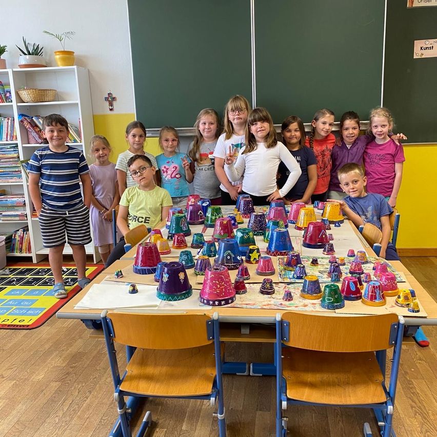 Eine Gruppe von Kindern steht um einen Tisch mit bemalten Bechern, in einem Klassenzimmer mit einer Tafel, Büchern und einem Teppich auf dem Boden.
