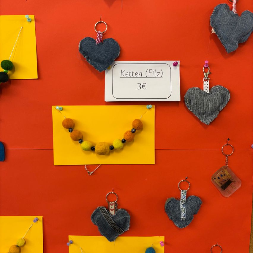 Eine rote Wand zeigt verschiedene Gegenstände, darunter Filzherzen, einen gelben Anhänger mit einer Perlenkette zum Preis von 3€ und weitere Accessoires.