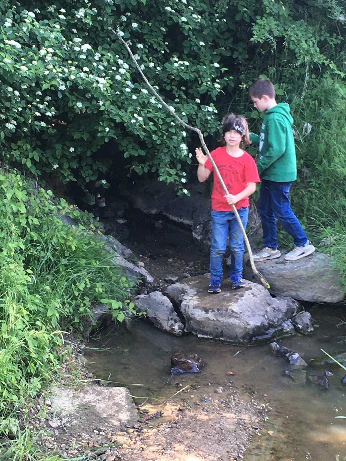 Zwei Kinder stehen auf Felsen über einem fließenden Bach, einer hält einen Stock. Der Junge in Rot winkt zur Kamera.