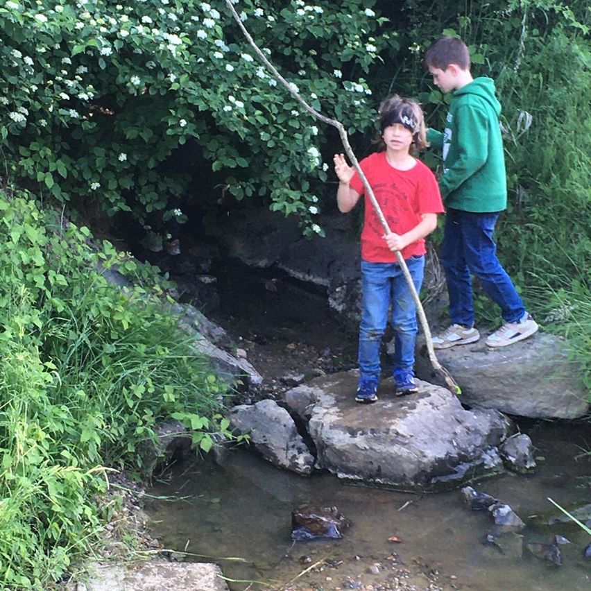 Zwei Kinder stehen auf Felsen über einem fließenden Bach, einer hält einen Stock. Der Junge in Rot winkt zur Kamera.