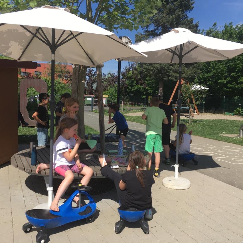 Eine Gruppe von Kindern versammelt sich um einen Tisch unter einem großen Schirm auf einem Spielplatz. Einige Kinder essen, während andere auf kleinen Fahrzeugen spielen. Im Hintergrund gibt es einen Rasenbereich und Bäume.