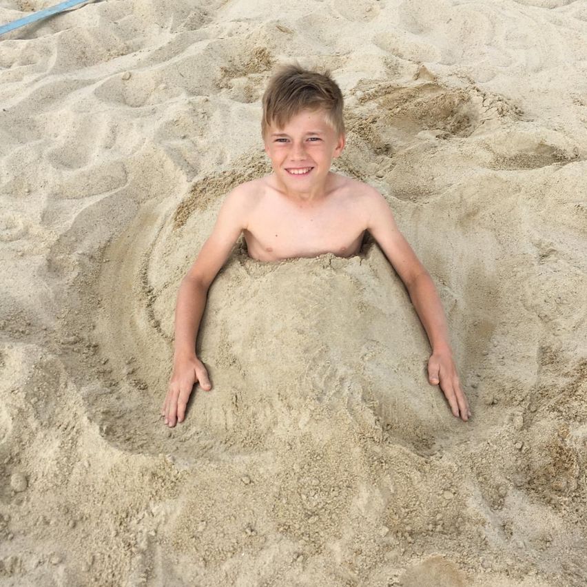 Ein junger Junge ist bis zum Hals im Sand am Strand begraben und lächelt in die Kamera.