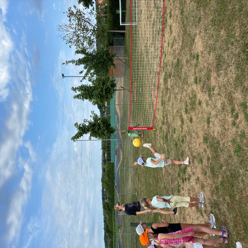 Eine Gruppe von Kindern spielt ein Spiel mit einem gelben Ball in einem Außenbereich mit einem Netz. Der Bereich ist von grünen Bäumen und einem klaren blauen Himmel umgeben.