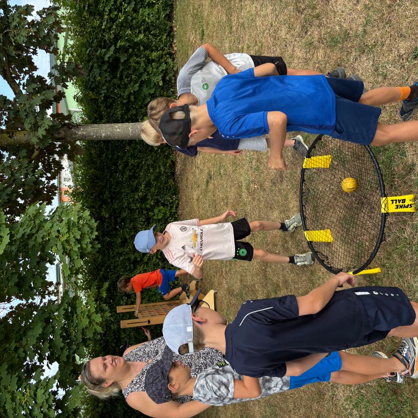 Mehrere Kinder spielen an einem sonnigen Tag draußen. Ein Kind hält einen Ball, während ein anderes vor einem Netz steht.