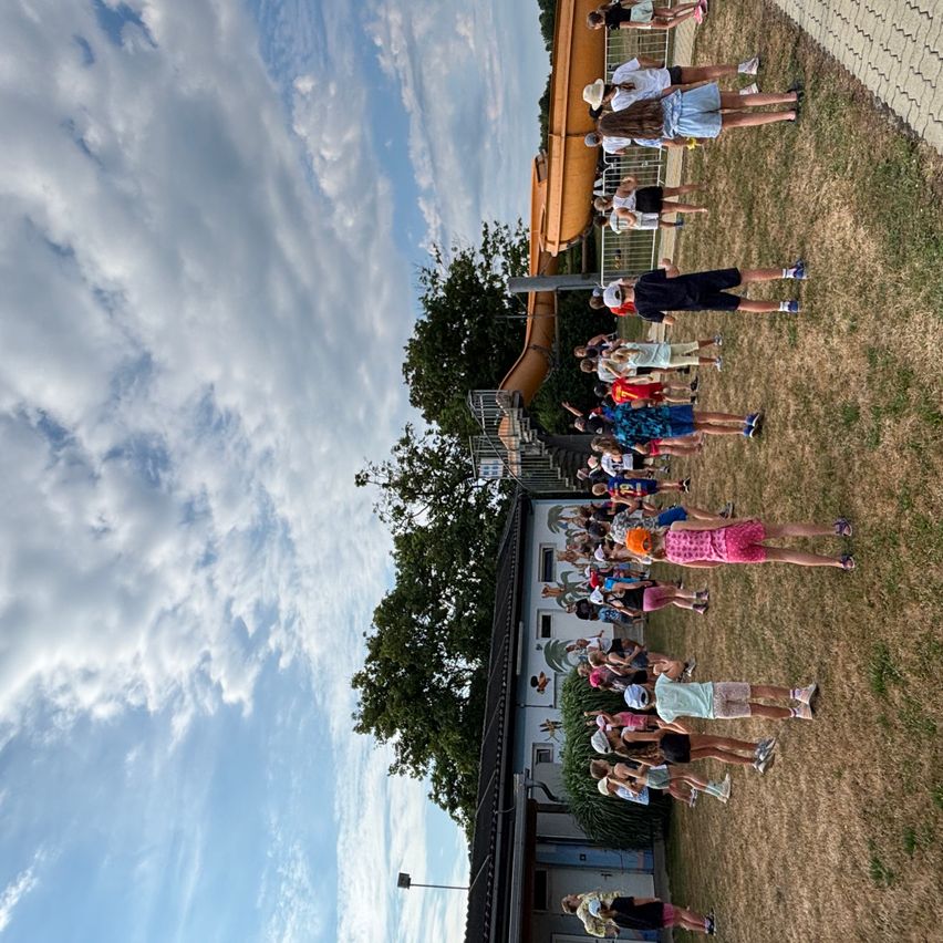 Eine Gruppe von Menschen steht auf einem Rasenfeld mit einer großen Rutsche hinter ihnen. Der Himmel ist blau mit weißen Wolken. Bäume und ein Gebäude sind im Hintergrund zu sehen.