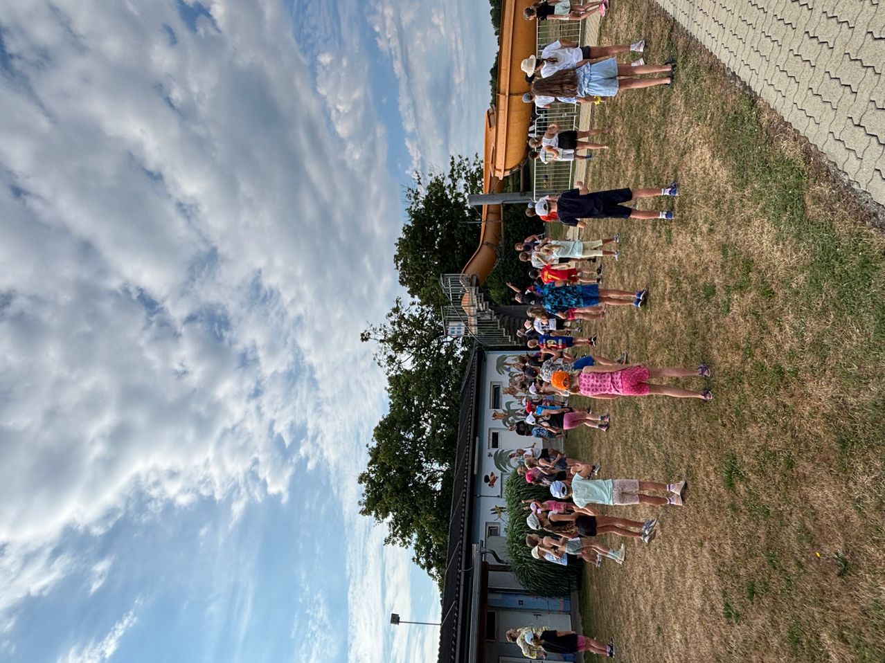 Eine Gruppe von Menschen steht auf einem Rasenfeld mit einer großen Rutsche hinter ihnen. Der Himmel ist blau mit weißen Wolken. Bäume und ein Gebäude sind im Hintergrund zu sehen.
