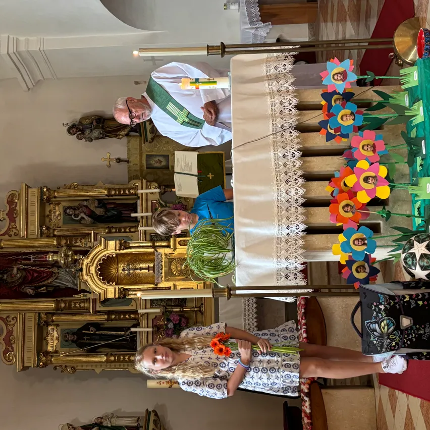 Ein Priester steht an einem Altar in einer Kirche, ein Junge kniet davor und ein Mädchen hält Blumen im Vordergrund. Bunte Blumen und eine Tasche sind auf dem Altar.