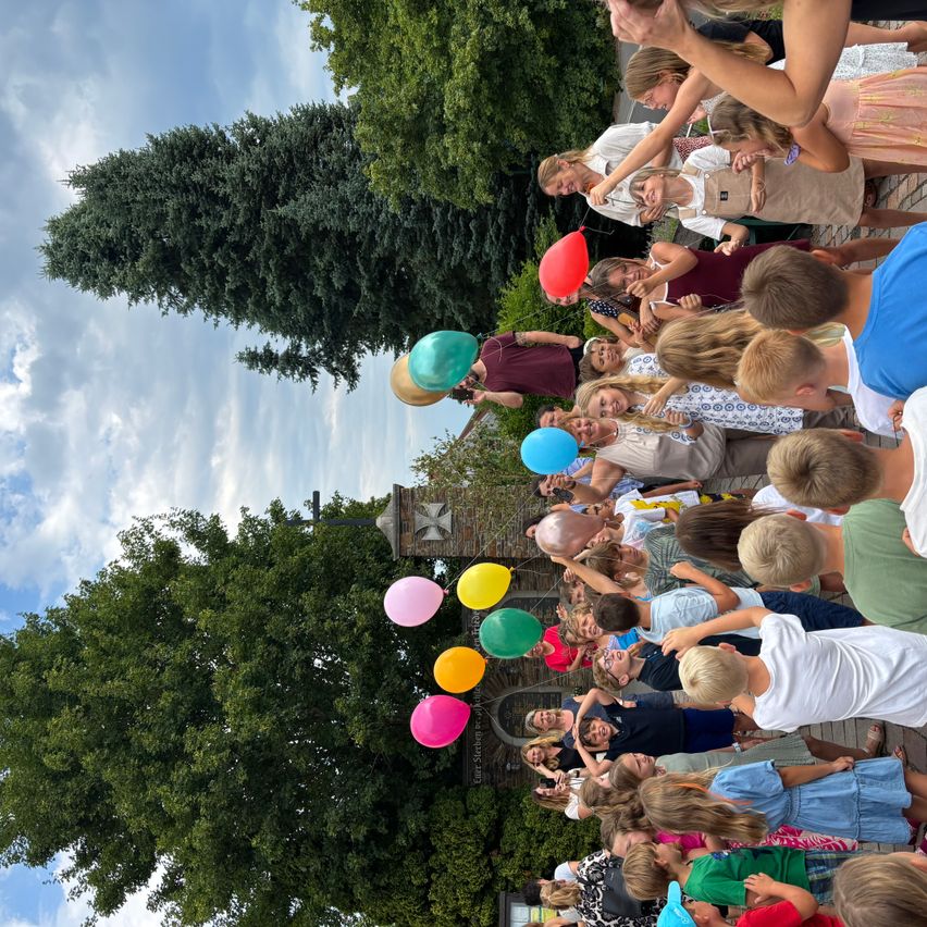Eine Gruppe von Kindern und Erwachsenen versammelt sich und hält bunte Luftballons. Sie stehen in einem Außenbereich mit Bäumen und blauem Himmel.