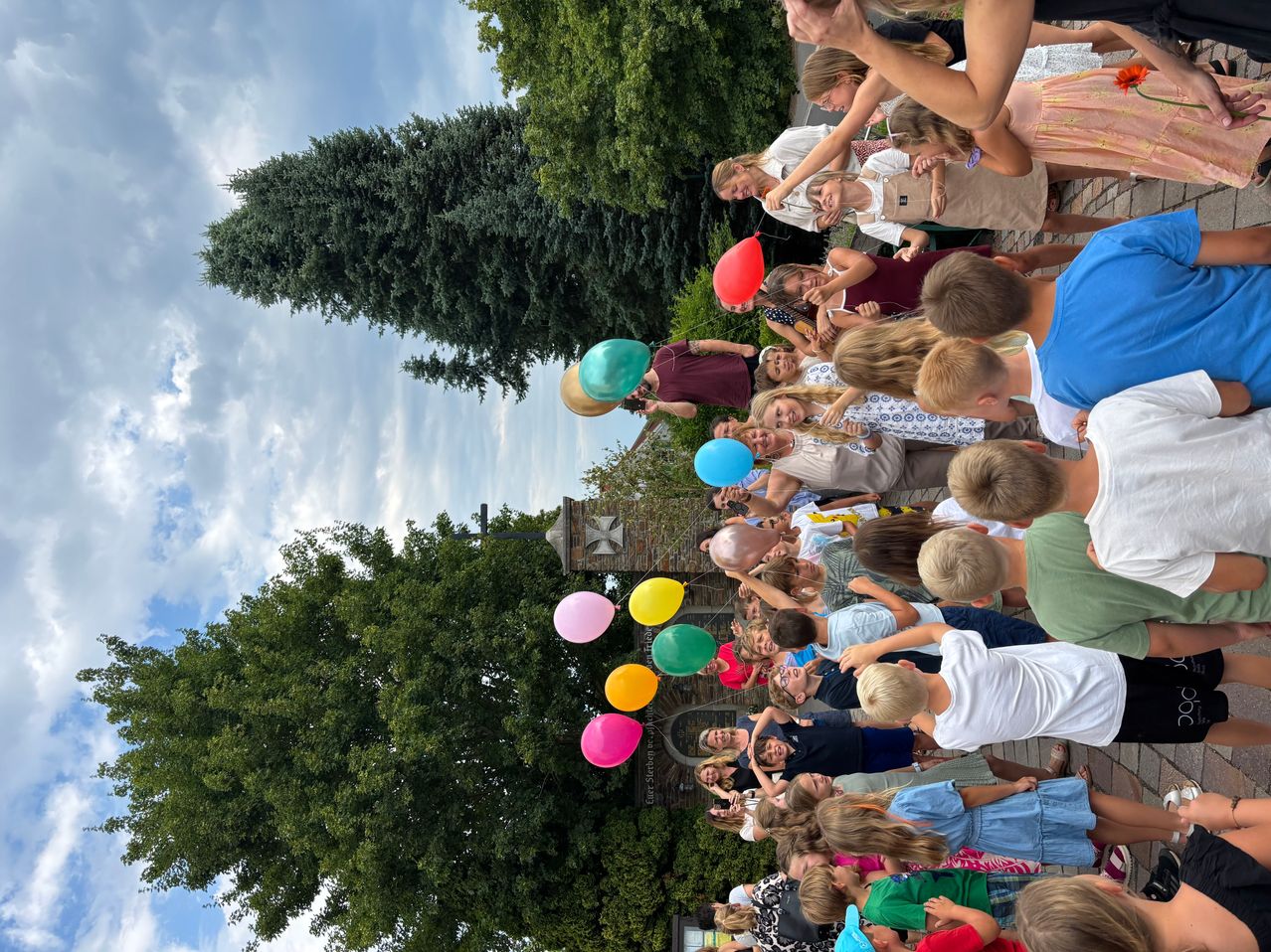 Eine Gruppe von Kindern und Erwachsenen versammelt sich und hält bunte Luftballons. Sie stehen in einem Außenbereich mit Bäumen und blauem Himmel.