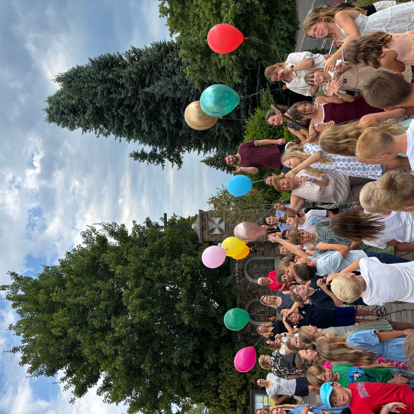 Eine Gruppe von Menschen versammelt sich im Freien und hält Luftballons in verschiedenen Farben. Sie stehen vor einem Baum und einem Steingebäude unter einem teilweise bewölkten Himmel.