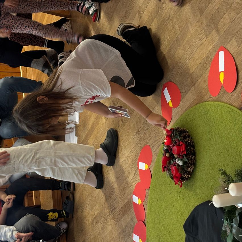 Mehrere Personen sitzen und hocken auf einem Holzboden. Ein Mädchen in Weiß greift nach einer mit Kerzen bestückten Girlande. Andere Dekorationen umgeben die Girlande.