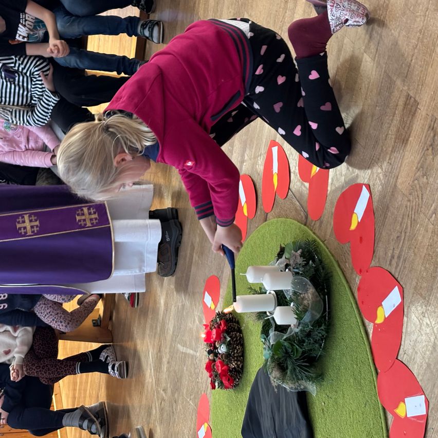 Eine Frau zündet Kerzen in einem geschmückten Kranz auf einem grünen Teppich an. Mehrere Menschen, darunter Kinder, sind um sie herum versammelt. Der Boden ist aus Holz.