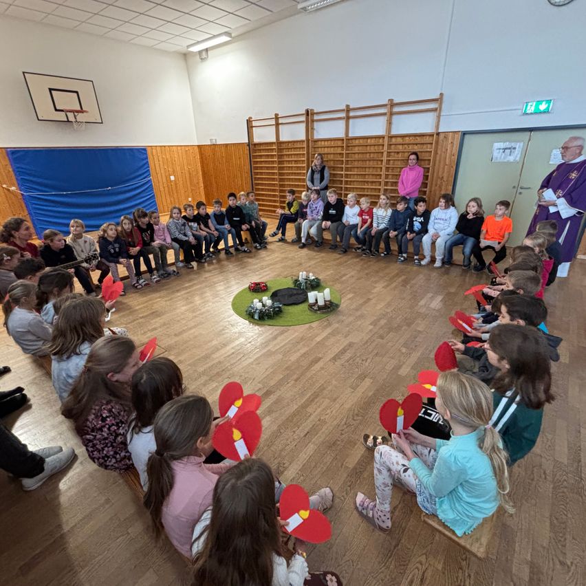 Eine Gruppe von Kindern sitzt in einem Kreis um einen Priester und hält rote Papierherzen in den Händen. Der Ort ist eine Turnhalle mit Holzböden und Wänden. Kerzen sind auf einem grünen Teppich in der Mitte entzündet.