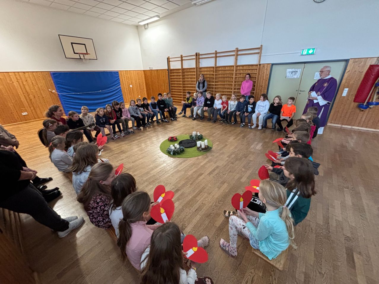 Eine Gruppe von Kindern sitzt in einem Kreis um einen Priester und hält rote Papierherzen in den Händen. Der Ort ist eine Turnhalle mit Holzböden und Wänden. Kerzen sind auf einem grünen Teppich in der Mitte entzündet.