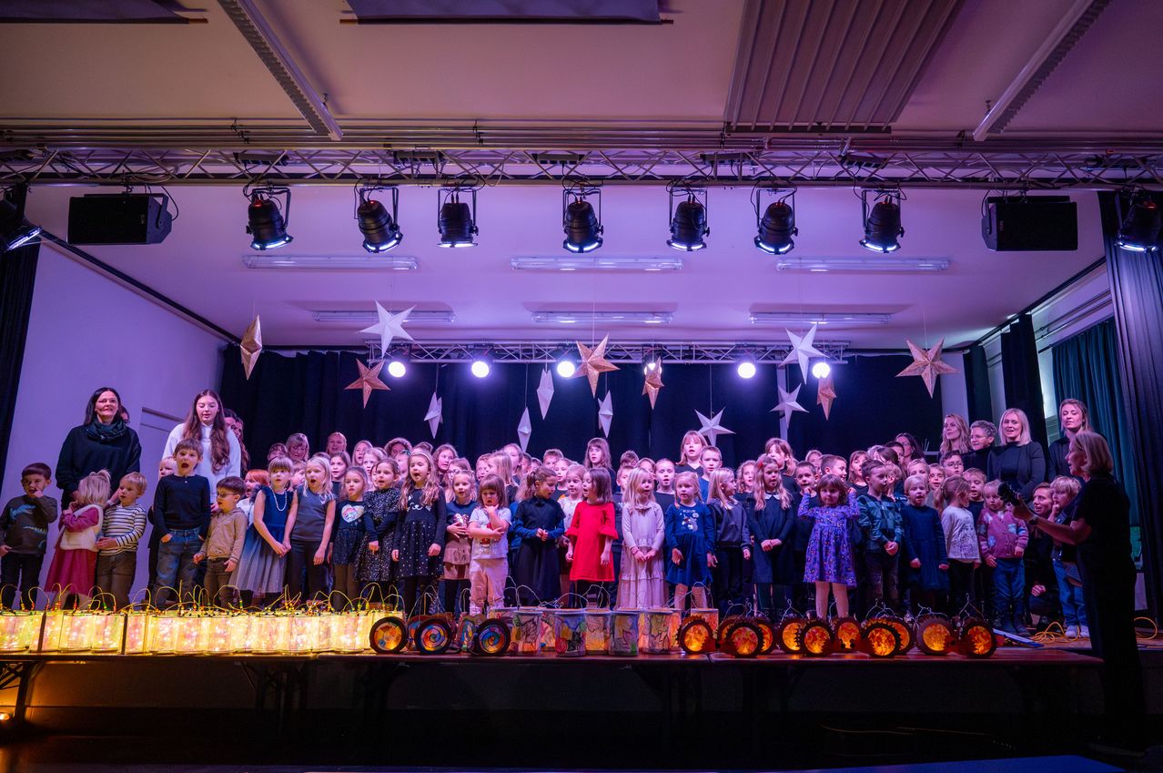 Eine Gruppe von Kindern steht auf einer Bühne und tritt auf. Sie halten Laternen, und Sterne hängen von der Decke. Lichter beleuchten die Bühne, und Erwachsene stehen im Hintergrund.