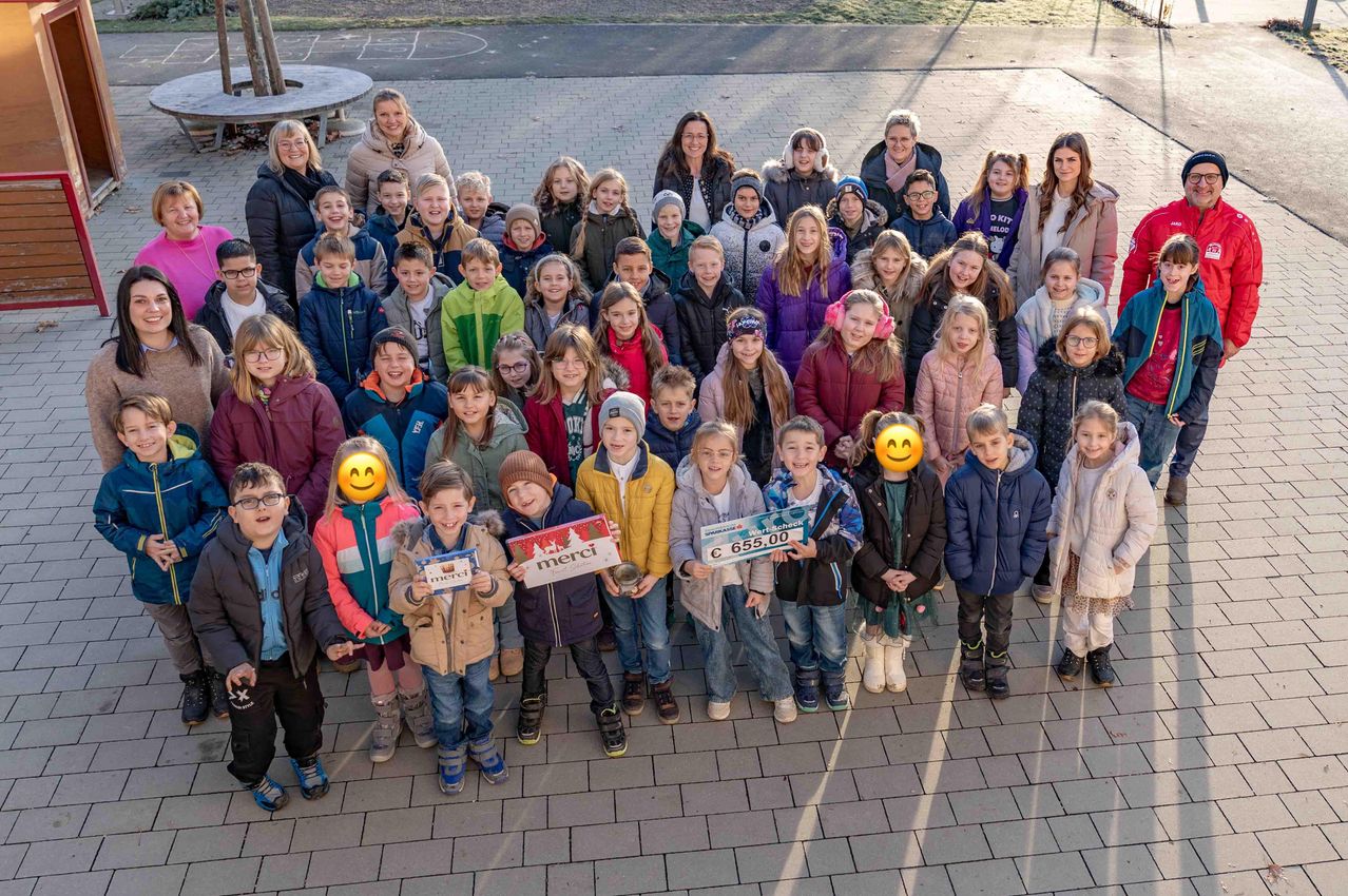 Eine Gruppe von Kindern und Erwachsenen, möglicherweise eine Schulklasse, posiert für ein Foto. Einige Kinder halten Boxen mit dem Wort 'Merci' darauf. Ein großes grünes Schild zeigt einen Spendenbetrag von 655,00 € an.