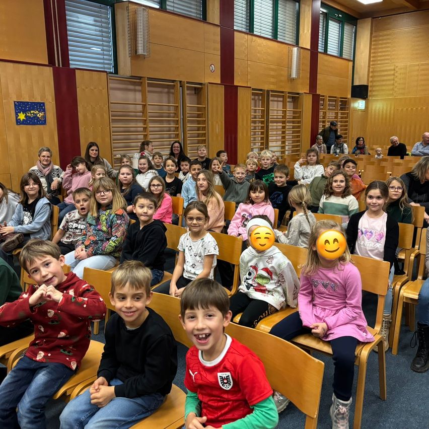 Ein Klassenzimmer voller Kinder ist in Reihen sitzend, einige tragen festliche Shirts und andere Winterkleidung. Zwei Kinder vorne haben Emoji-Aufkleber auf den Gesichtern.