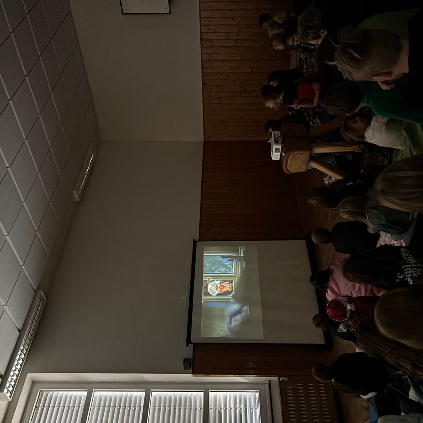 Eine Gruppe von Kindern ist drinnen versammelt und schaut auf eine Projektionsleinwand. Der Projektor zeigt ein Bild an. Der Raum hat Holzwände und weiße Fliesen.