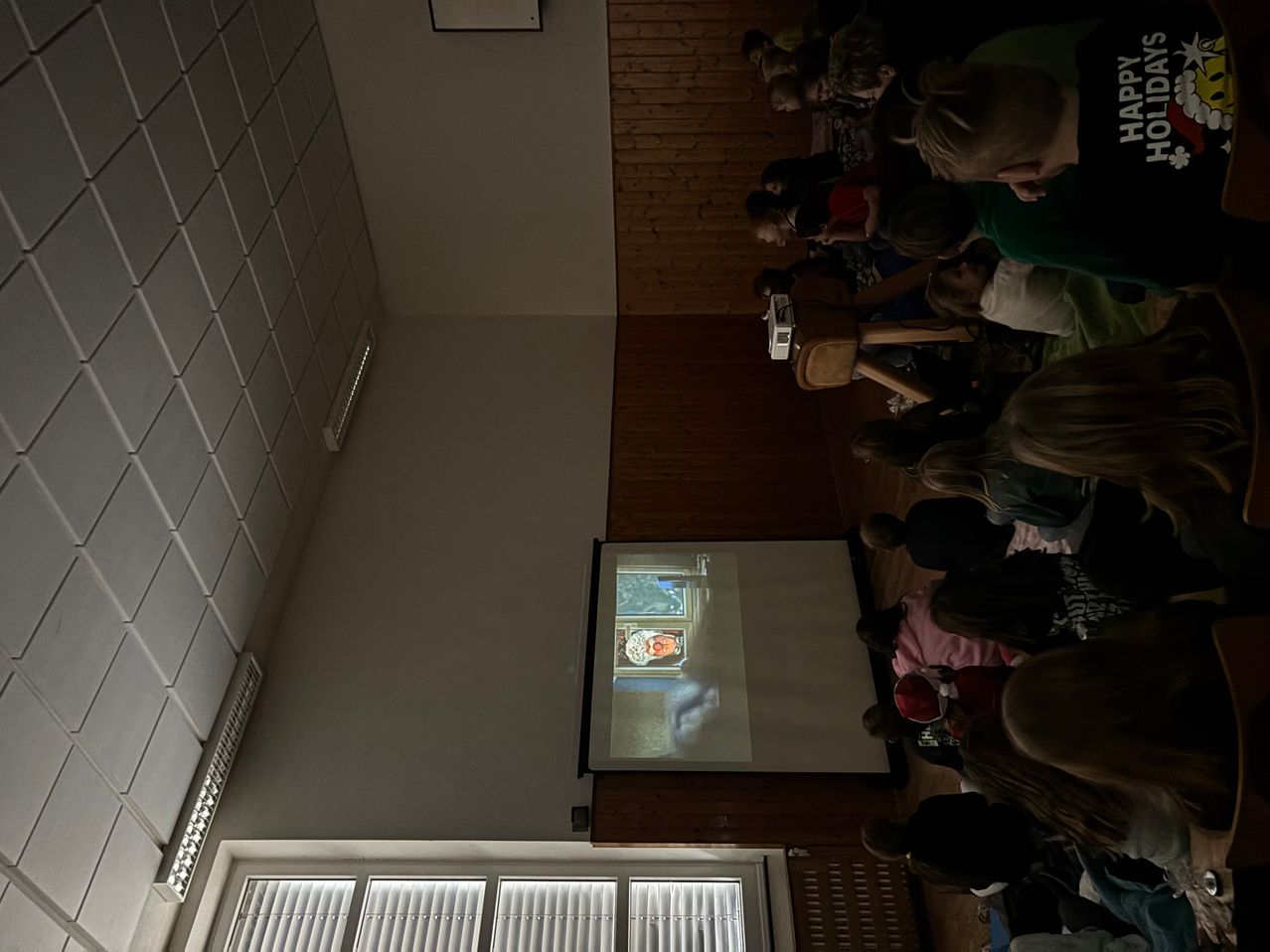 Eine Gruppe von Kindern ist drinnen versammelt und schaut auf eine Projektionsleinwand. Der Projektor zeigt ein Bild an. Der Raum hat Holzwände und weiße Fliesen.