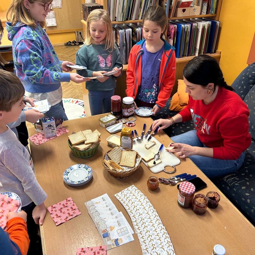 Eine Gruppe von Kindern und einer Frau versammelt sich um einen Tisch. Es gibt Sandwiches, Brot und Gläser mit Aufstrichen auf dem Tisch. Die Frau sitzt und scheint Essen zu servieren.