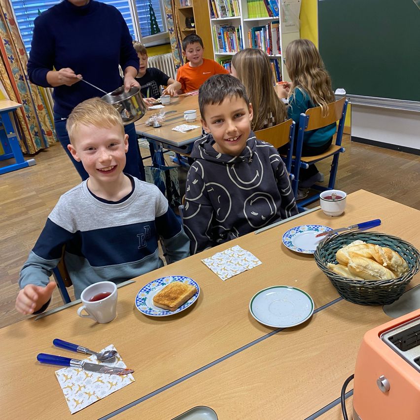 Kinder sitzen an einem Tisch in einem Klassenzimmer und lächeln. Ein Junge hält eine Tasse mit Getränk. Ein anderer Junge sitzt neben ihm mit einem Teller Toast. Eine Lehrerin steht hinter ihnen.