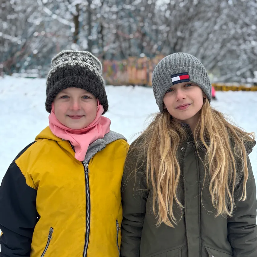 Zwei Mädchen posieren im Schnee, beide in Wintermänteln und Mützen. Das Mädchen auf der linken Seite trägt einen gelben Mantel und einen rosa Schal, während das Mädchen auf der rechten Seite einen grünen Mantel und eine Tommy Hilfiger-Mütze trägt.