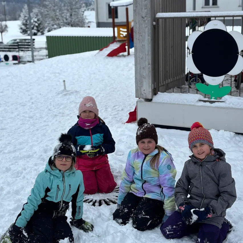 Vier Kinder sitzen im Schnee, eingehüllt in warme Winterkleidung. Hinter ihnen befindet sich ein großes Blumendesign auf einem Balkon, und im Hintergrund sind eine Rutsche und Spielgeräte zu sehen. Der schneebedeckte Boden erstreckt sich in die Ferne, wobei Bäume und ein Gebäude sichtbar sind.