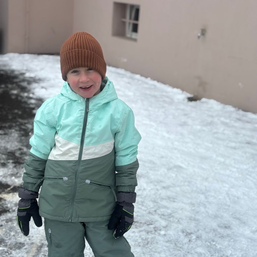 Ein junger Junge in einer türkisfarbenen und grünen Winterjacke, der lächelnd im Schnee steht. Er trägt eine braune Mütze und schwarze Handschuhe. Hinter ihm ist ein Gebäude mit einem Fenster zu sehen.