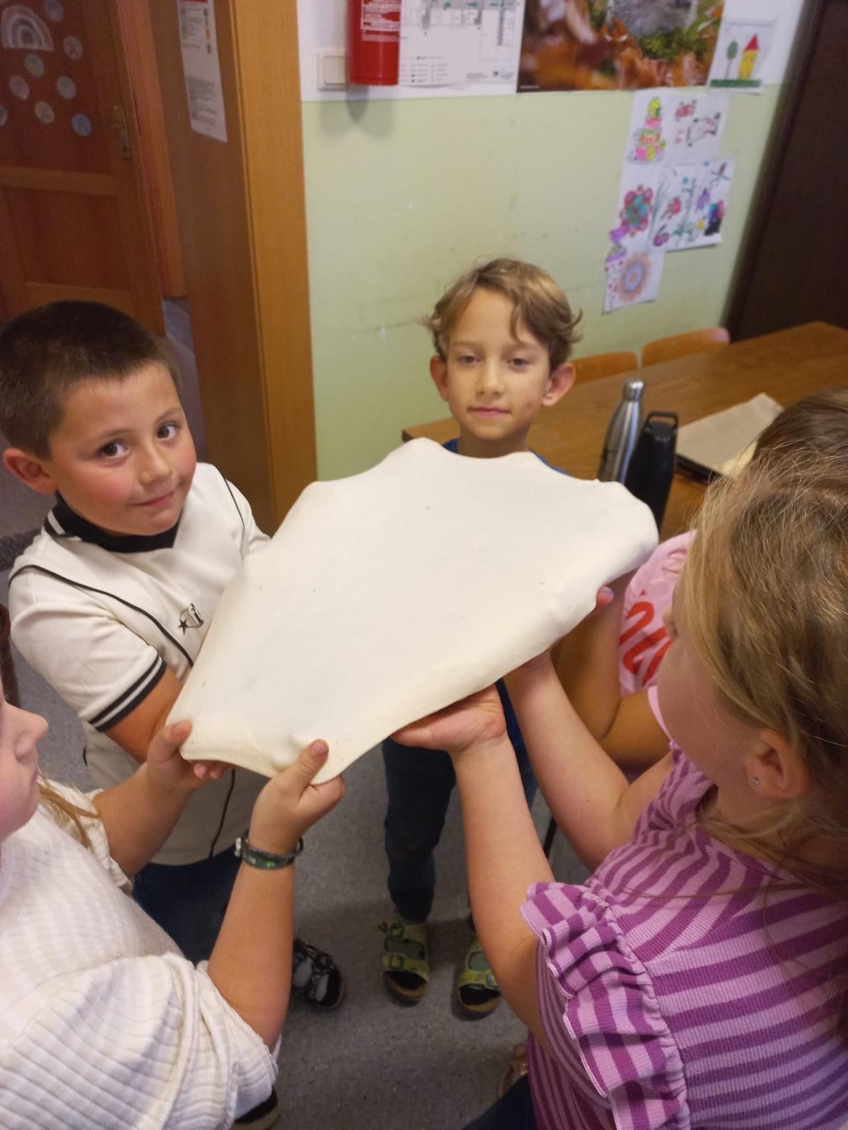 Vier Kinder stehen in einem Raum und halten ein großes Stück Teig. Zwei Mädchen halten den Teig und zwei Jungen schauen zu. Ein Tisch mit Gegenständen steht hinter ihnen.