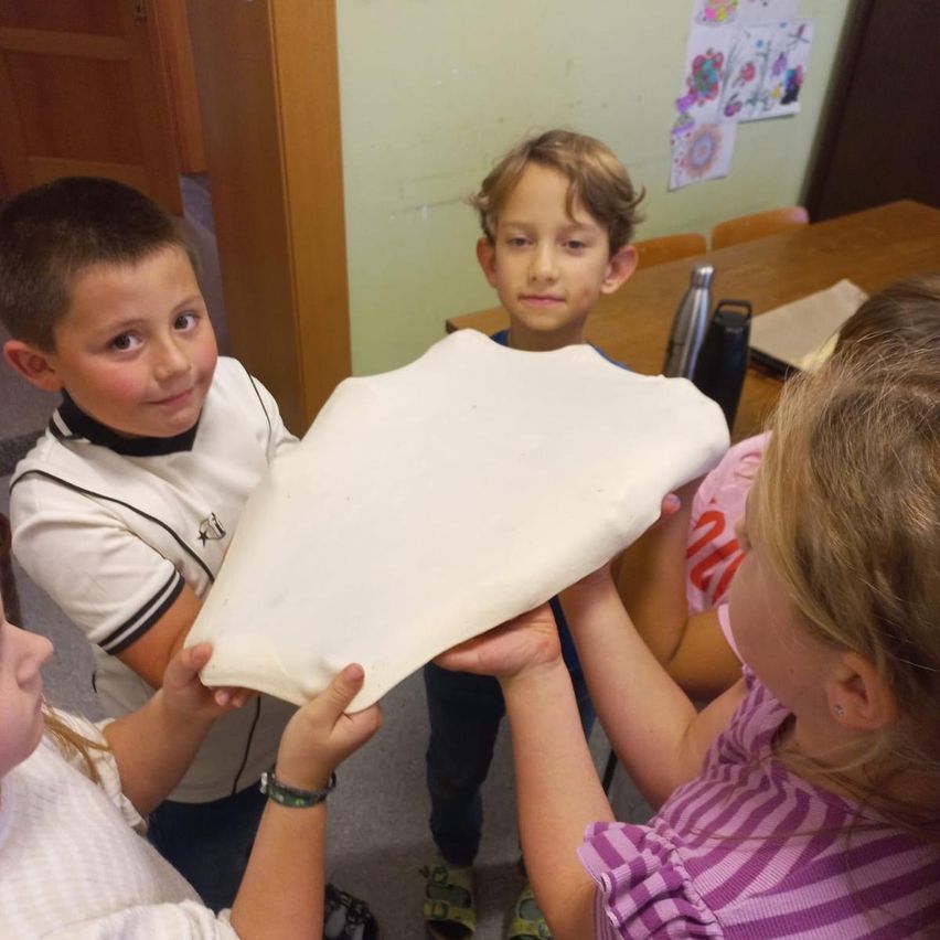 Vier Kinder stehen in einem Raum und halten ein großes Stück Teig. Zwei Mädchen halten den Teig und zwei Jungen schauen zu. Ein Tisch mit Gegenständen steht hinter ihnen.