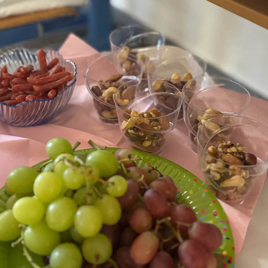 Ein Tisch mit einem rosa Tuch hat einen Teller mit Trauben, eine Schüssel mit Würstchen und Bechern mit Nüssen gefüllt.