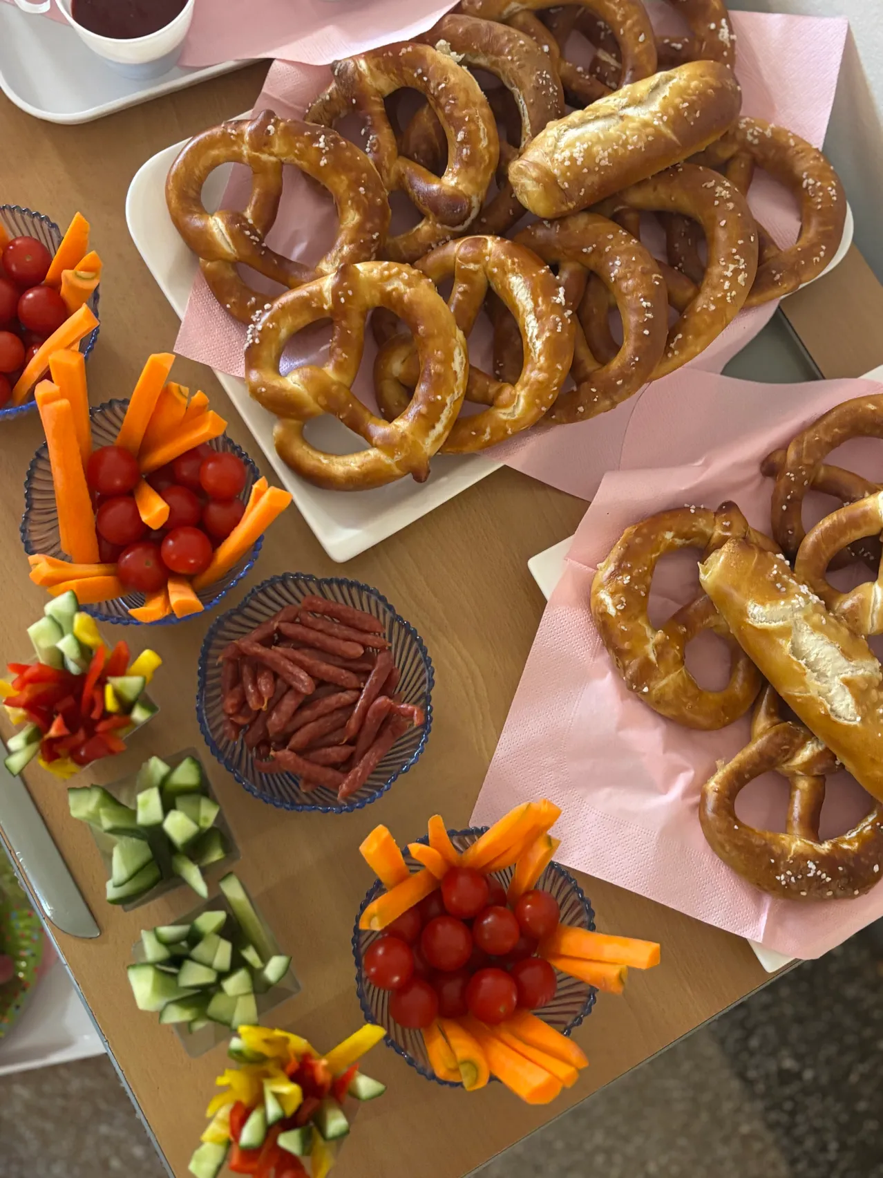 Ein Holztisch mit verschiedenen Lebensmitteln, darunter Brezeln, Karotten, Tomaten und Würstchen, in Schüsseln auf rosa Servietten angeordnet.