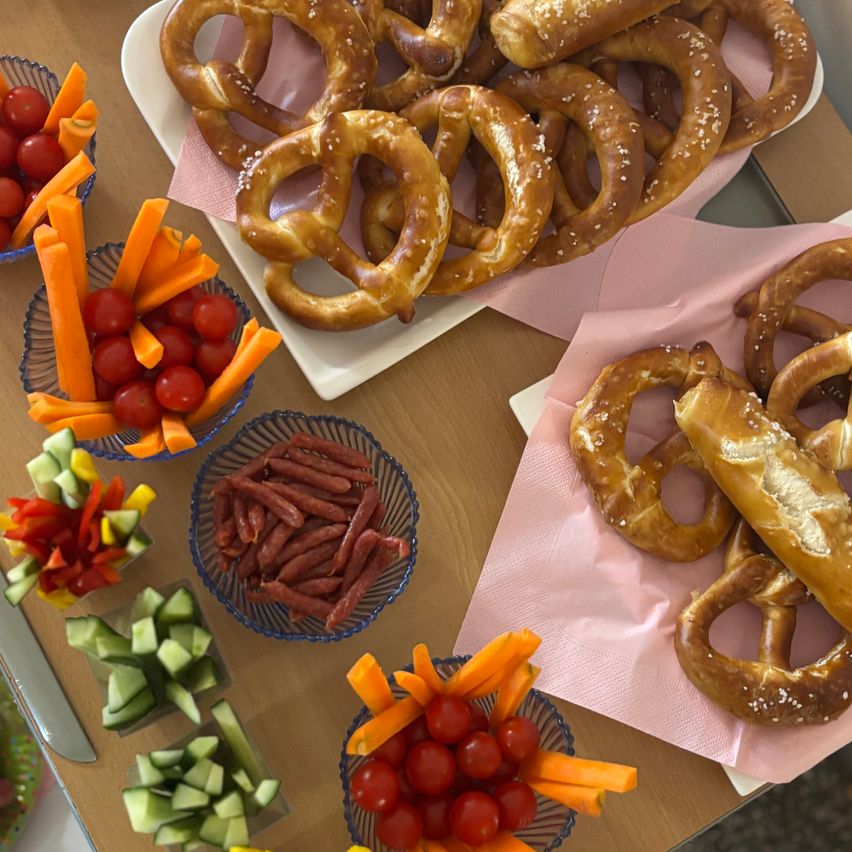 Ein Holztisch mit verschiedenen Lebensmitteln, darunter Brezeln, Karotten, Tomaten und Würstchen, in Schüsseln auf rosa Servietten angeordnet.