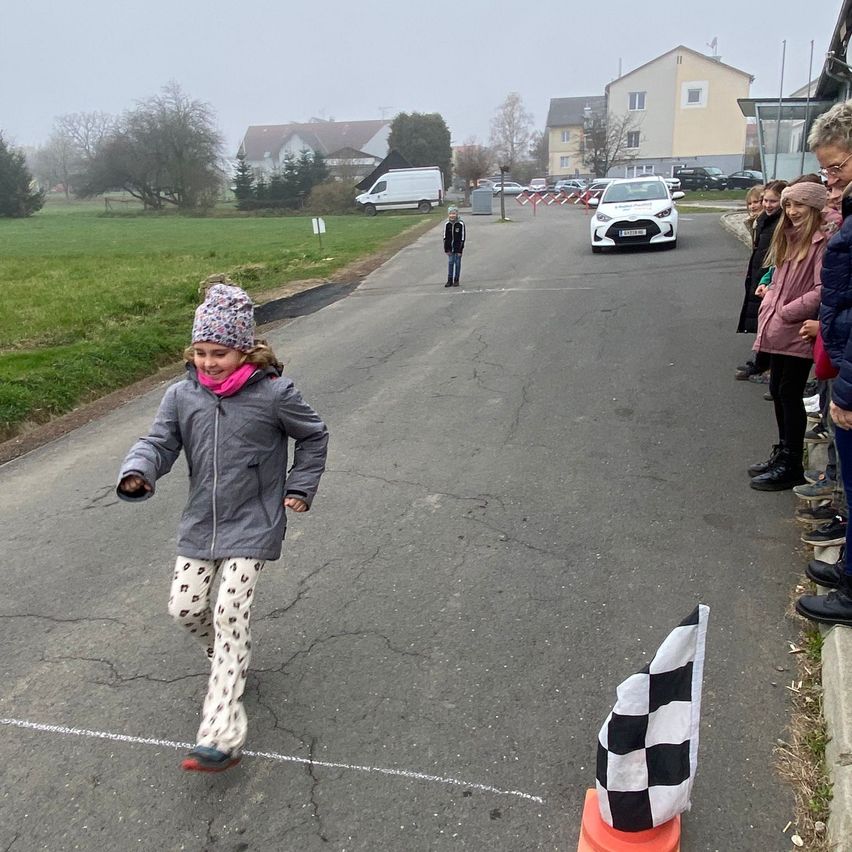 Ein junges Mädchen geht auf einer Straße mit einer Schachbrettfahne in der Nähe. Zuschauer beobachten sie von der Seite. Im Hintergrund steht ein geparktes weißes Auto und mehrere Häuser.