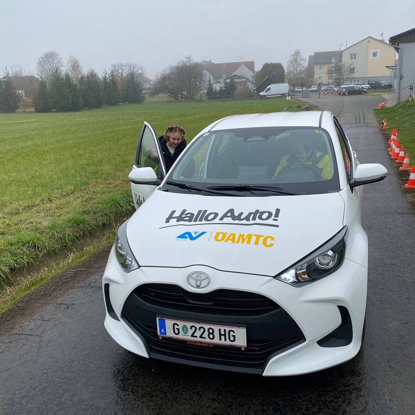 Ein weißer Toyota Yaris mit Hallo Auto und DAMTC-Text auf der Motorhaube steht auf einer Straße mit einem Grasfeld und Verkehrskegeln. Eine Frau steht neben der offenen Tür.