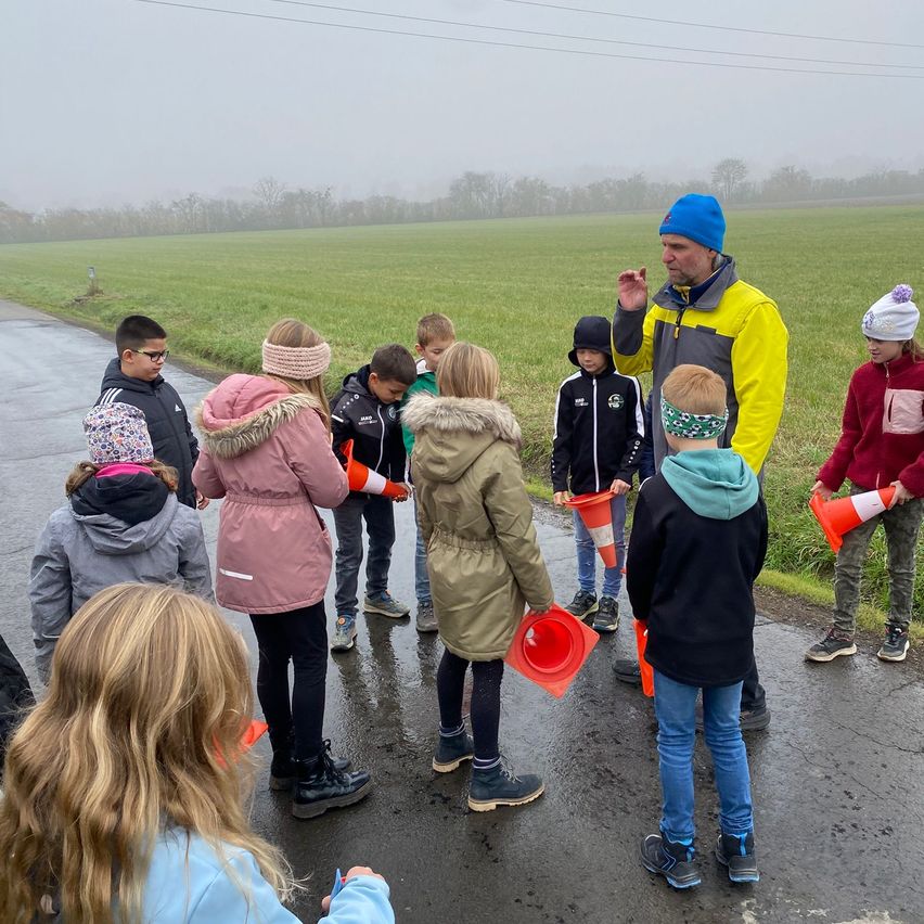 Eine Gruppe von Kindern und Erwachsenen steht auf einer nassen Straße, bekleidet mit Jacken und Mützen. Sie halten orangefarbene Verkehrskegel in den Händen, und ein Mann in einer gelben Jacke gibt ihnen Anweisungen. Im Hintergrund befindet sich ein grünes Feld mit Nebel.