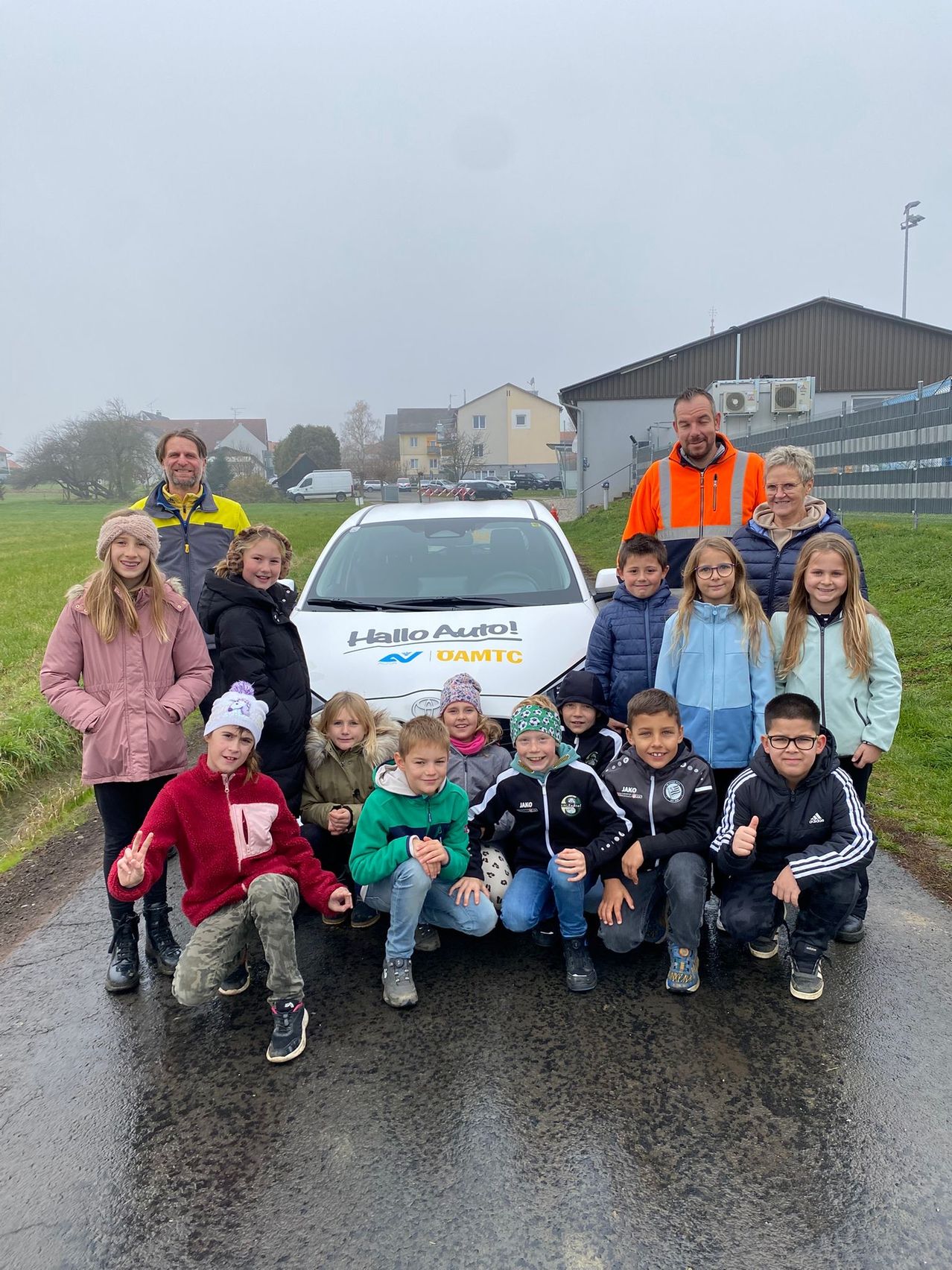 Eine Gruppe von Kindern und Erwachsenen posiert vor einem weißen Elektroauto mit der Aufschrift 'Hello Auto!'. Sie alle lächeln und scheinen für ein Foto zu posieren.