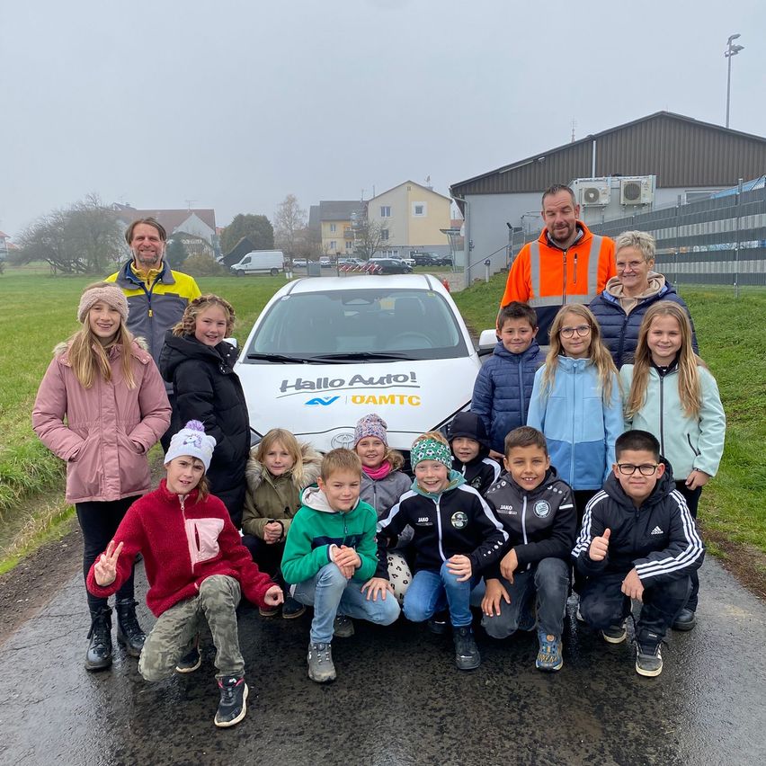Eine Gruppe von Kindern und Erwachsenen posiert vor einem weißen Elektroauto mit der Aufschrift 'Hello Auto!'. Sie alle lächeln und scheinen für ein Foto zu posieren.