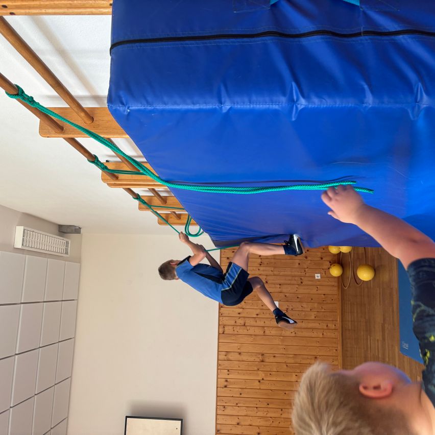 Ein Mann hängt an einer Seilleiter, die an einem großen blauen Kissen an der Decke befestigt ist, während ein anderer zusieht. Der Raum hat Holzwände und gelbe Bälle.