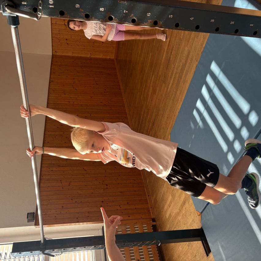 Ein junges Kind führt einen Klimmzug an einer Stange in einem Fitnessstudio durch, während ein anderes Kind von hinten zuschaut.