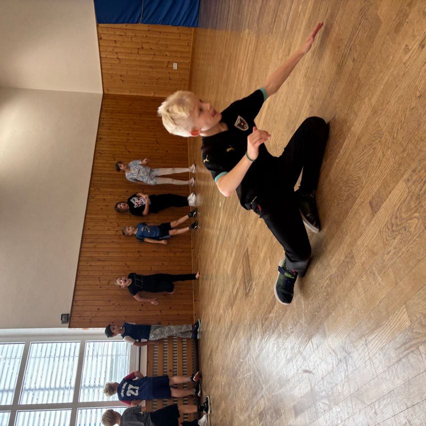 Eine Gruppe von Kindern übt Kampfkunst auf einem Holzboden mit einer Holzwand im Hintergrund. Ein Kind in Schwarz ist in einer Kampfhaltung.