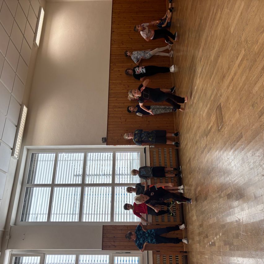 Eine Gruppe von Kindern übt Kampfsport in einer Turnhalle. Sie befinden sich in verschiedenen Haltungen. Der Raum hat Holzböden und weiße Wände.