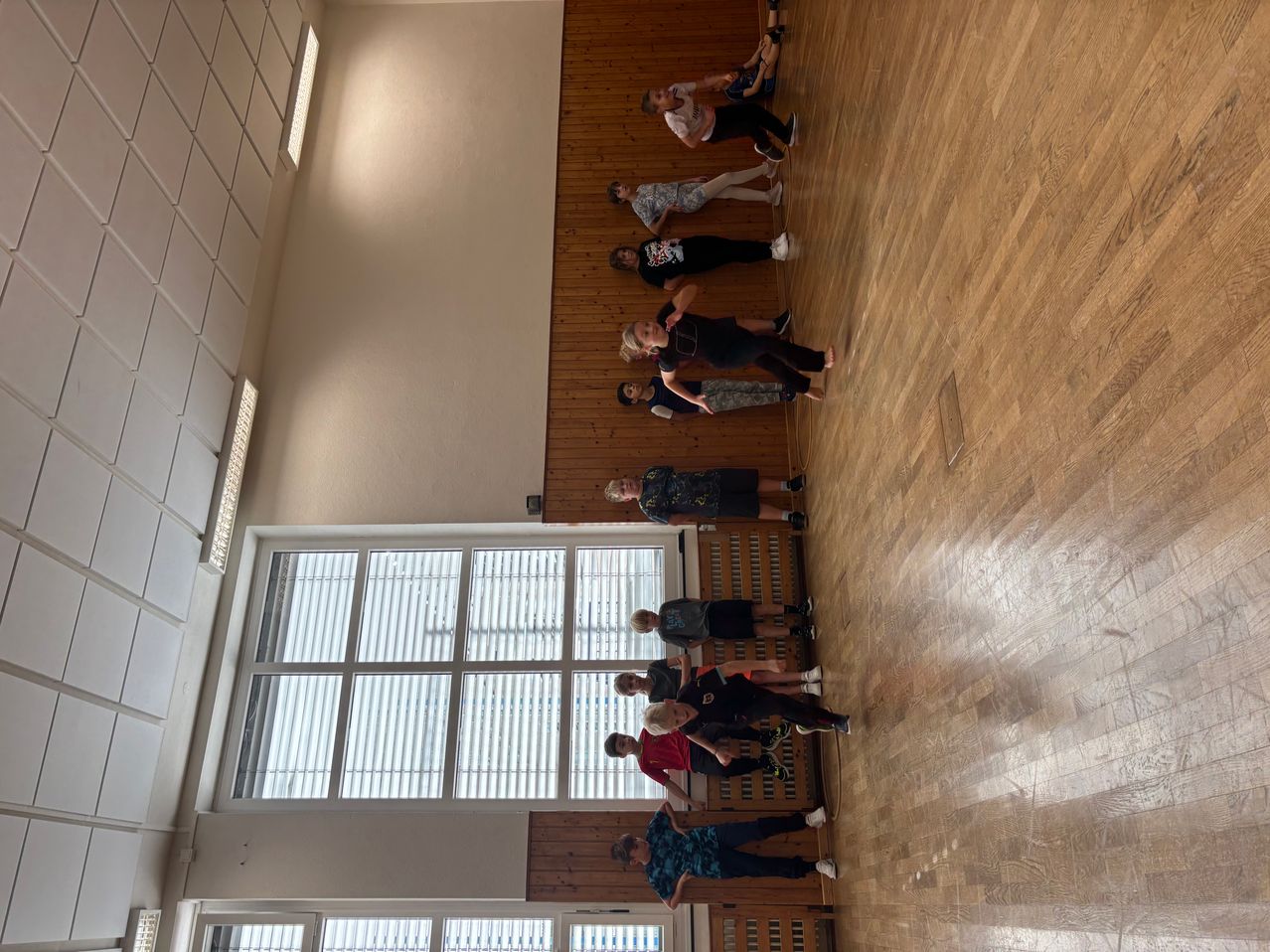 Eine Gruppe von Kindern übt Kampfsport in einer Turnhalle. Sie befinden sich in verschiedenen Haltungen. Der Raum hat Holzböden und weiße Wände.