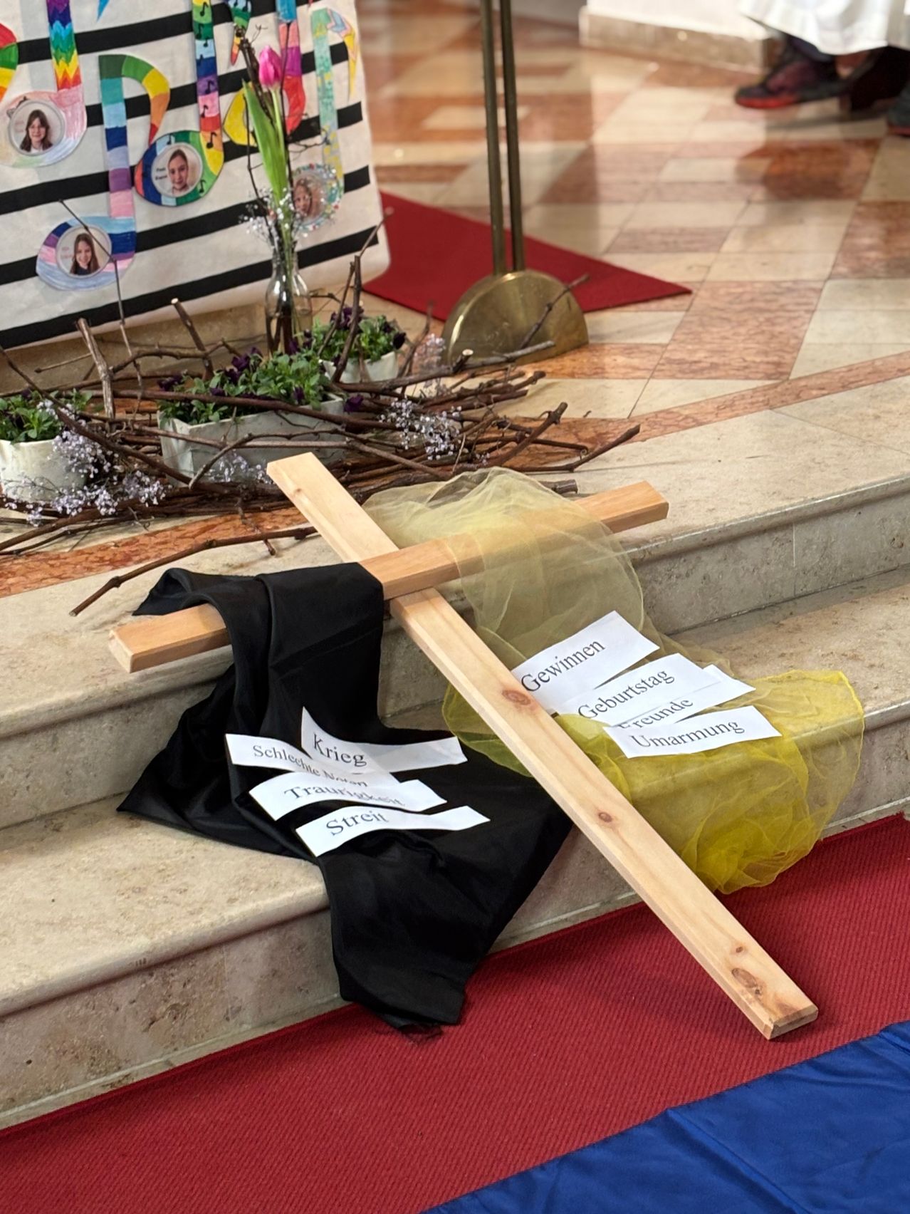 Ein hölzerne Kreuz mit schwarzem Tuch liegt auf einer Kirchenstufe. Daneben sind gelber Stoff und kleine Pflanzen zu sehen. Das Kreuz hat Etiketten wie 'Gewinnen', 'Geburtstag' und 'Umarmung'. Dahinter befindet sich ein roter Teppich und ein weißer Stoff mit Bildern ziert den Hintergrund.